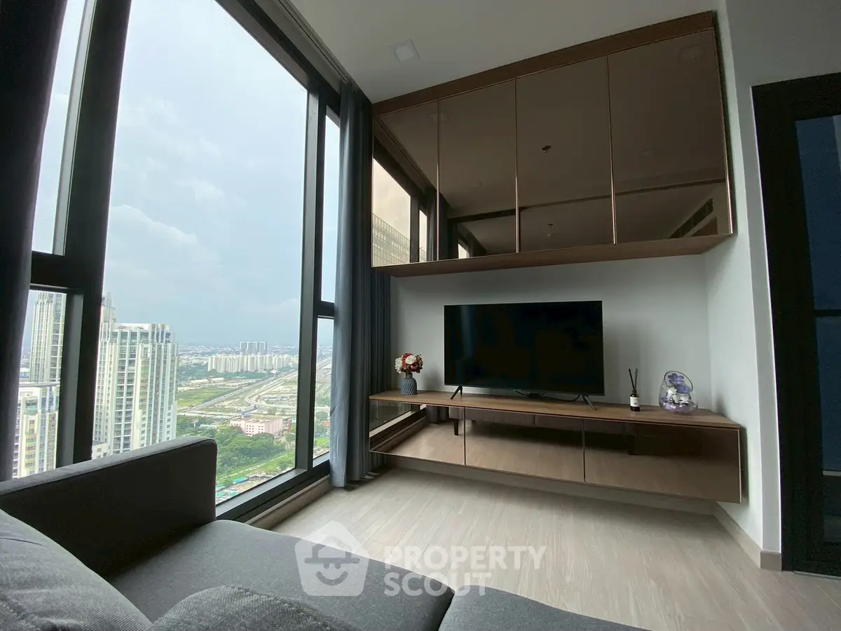 Modern living room with large windows and city view, featuring sleek furniture and a mounted TV.