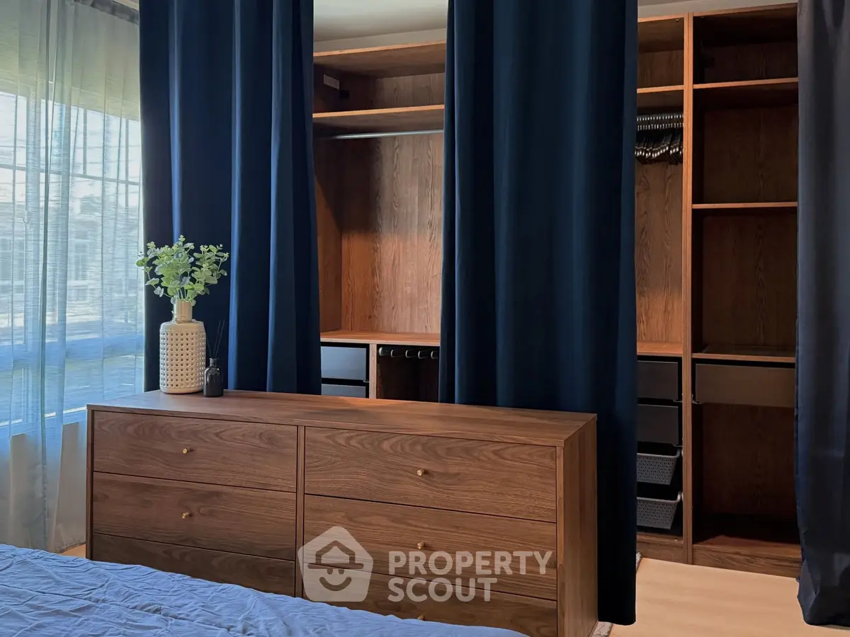 Spacious bedroom with elegant wooden dresser and open closet, featuring dark curtains and natural light.
