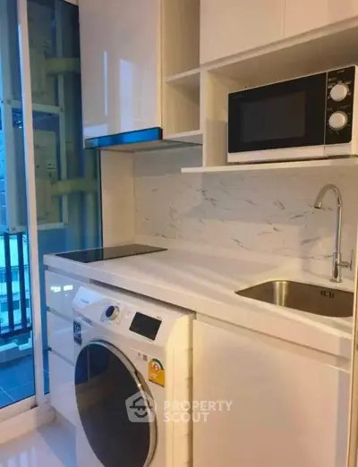 Modern compact kitchen with washing machine and microwave, sleek white cabinetry, and balcony view.