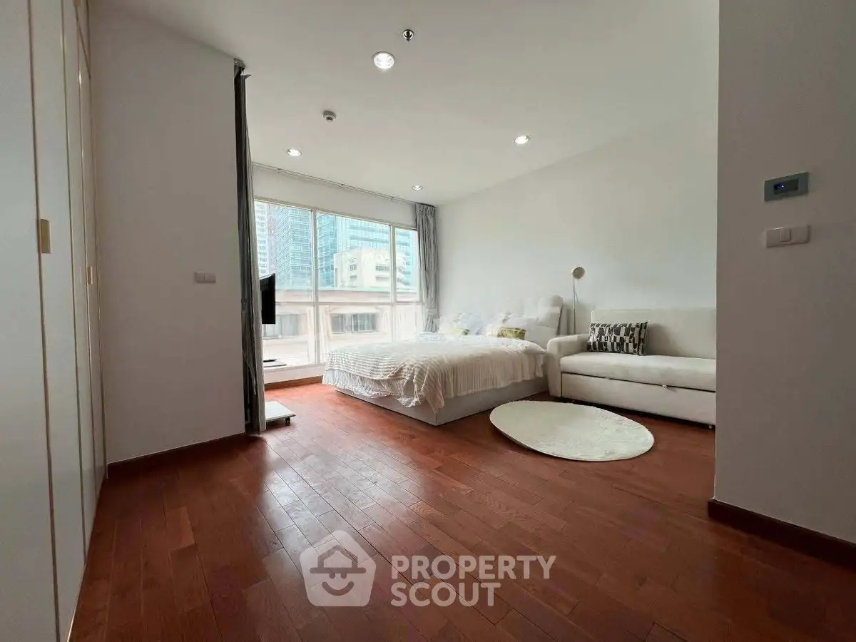 Spacious bedroom with wooden flooring and large window offering city views, featuring a cozy bed and modern decor.
