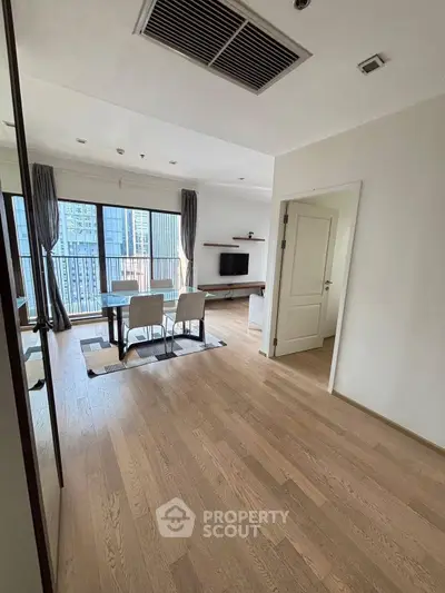 Modern living room with wooden flooring and city view, featuring a dining area and wall-mounted TV.