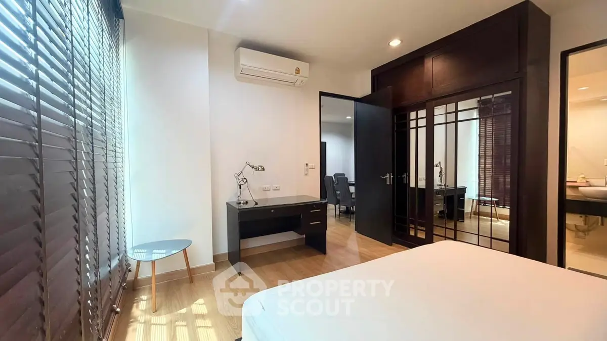 Spacious bedroom with modern furnishings and natural light, featuring a desk and large windows with blinds.
