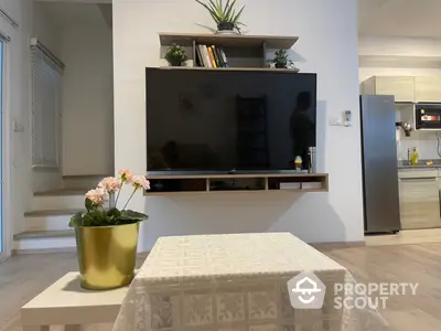 Modern living room with large flat-screen TV, sleek shelving, and an open-plan layout leading to a well-equipped kitchen.