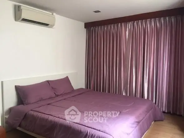Cozy bedroom with purple bedding and curtains, featuring air conditioning and wooden flooring.