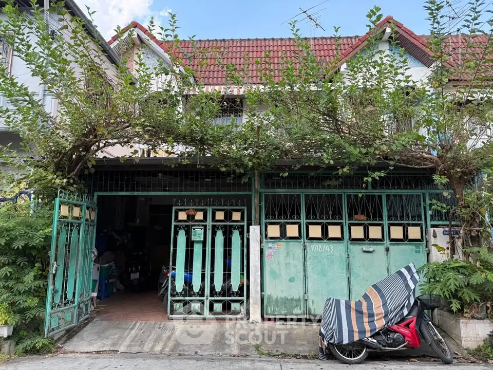 Charming townhouse with lush greenery and secure gated entrance, featuring a covered motorcycle parking area.