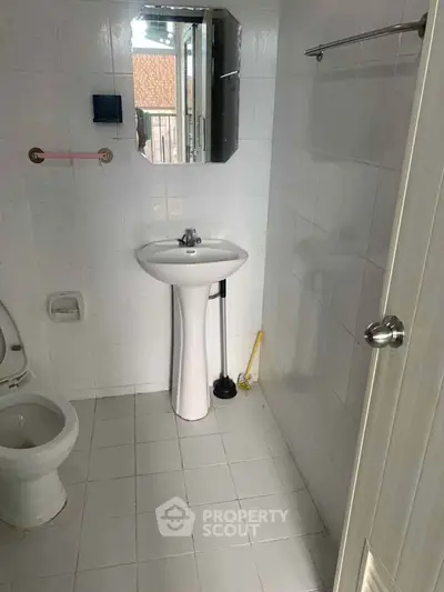 Compact bathroom with white tiles, pedestal sink, and toilet, featuring a small mirror and window for natural light.