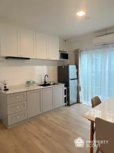 Modern kitchen with sleek cabinetry and natural light, featuring a fridge and dining area.