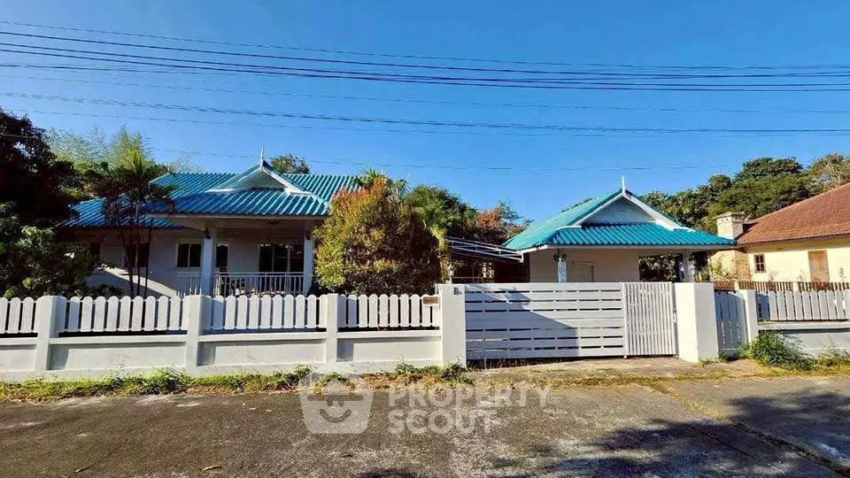 Charming blue-roofed house with white picket fence and lush greenery, perfect for serene living.