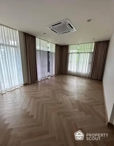 Spacious empty room with elegant herringbone wood flooring and large windows.