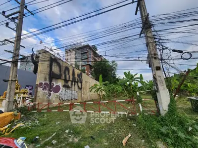 Urban land plot with potential for development, featuring a backdrop of a multi-story building and lush greenery.