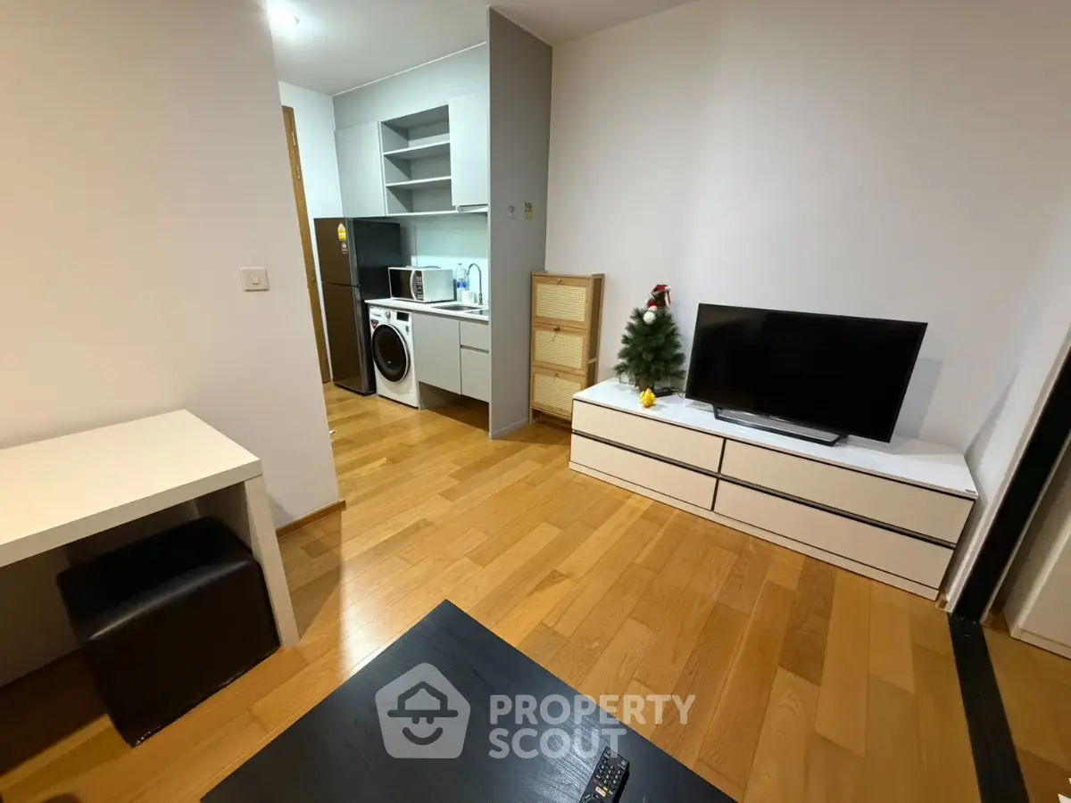 Modern living room with wooden flooring, TV unit, and compact kitchen area featuring washing machine and microwave.