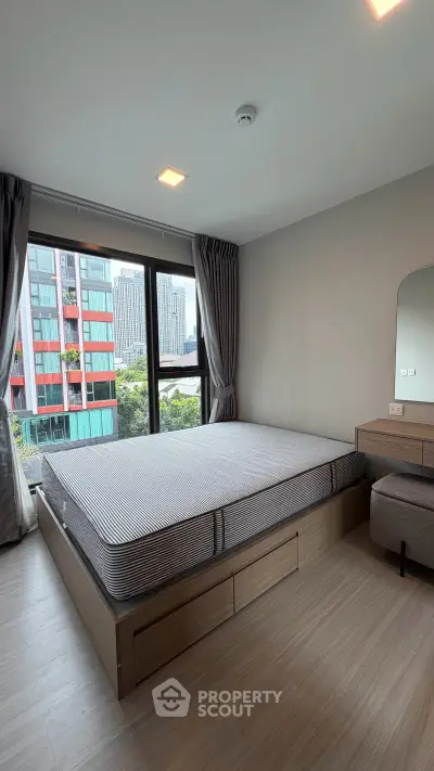 Modern bedroom with large window offering city view, featuring sleek furniture and neutral tones.