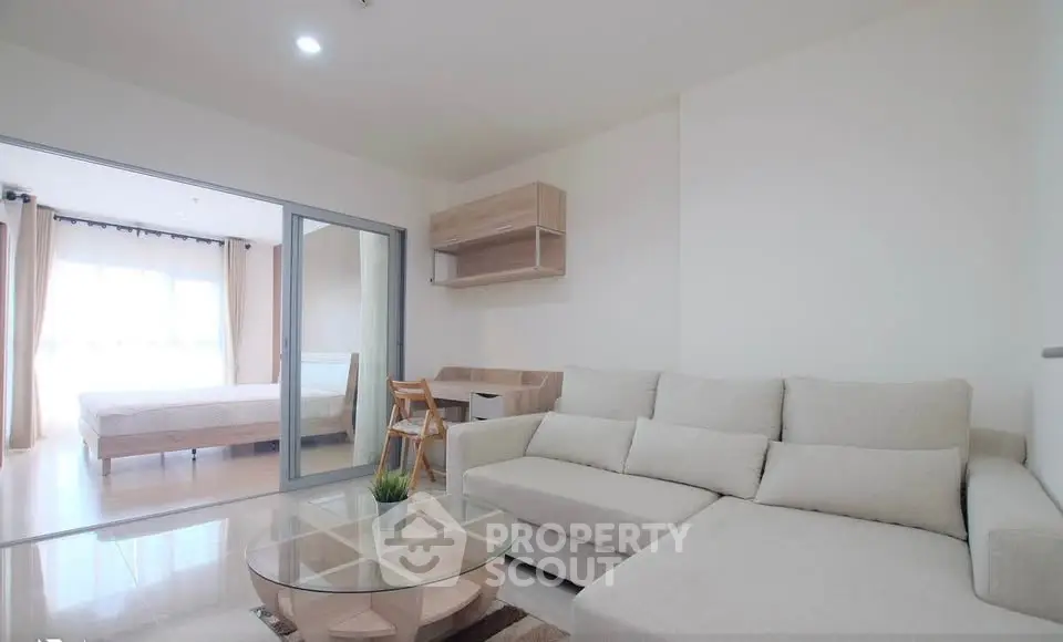 Modern living room with beige sectional sofa and glass coffee table, adjacent to a bright bedroom.