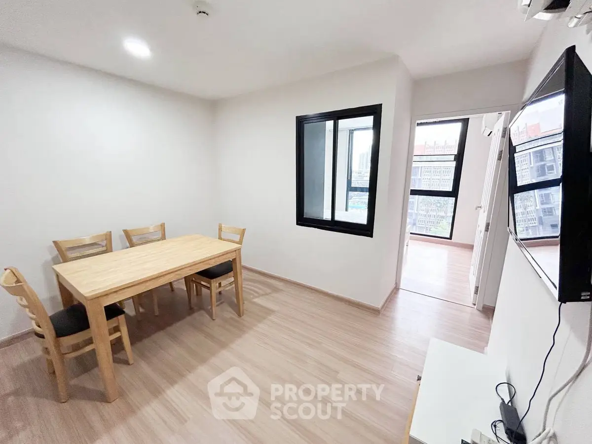 Modern dining area with wooden table and chairs, adjacent to a bright room with large windows.