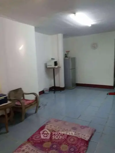 Spacious living area with tiled flooring and minimal furnishings, featuring a fridge and microwave.