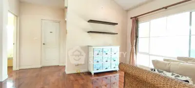 Bright living room with wooden floors and large window, featuring cozy seating and decorative shelves.