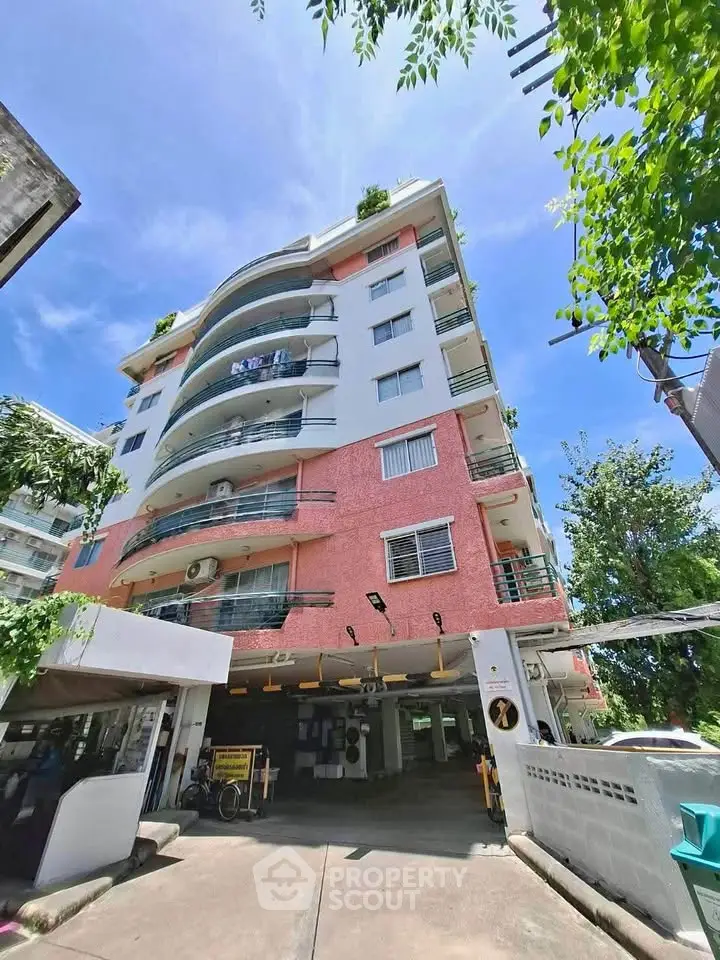 Modern multi-story residential building with balconies and parking area, surrounded by greenery under a clear blue sky.
