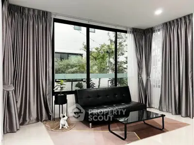 Modern living room with sleek black sofa, glass coffee table, and large windows with silver curtains.