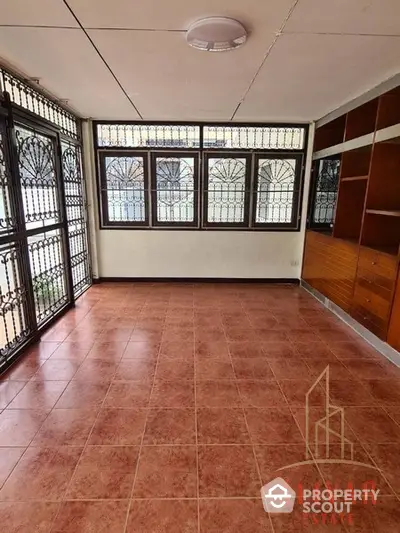 Spacious and well-lit room with terracotta tiled flooring, large windows with decorative grills, and built-in wooden shelving, ideal for a comfortable living space.