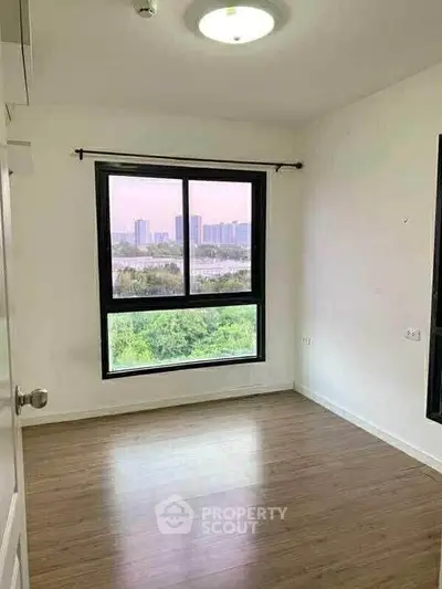 Bright empty room with large window offering cityscape view and wooden flooring.