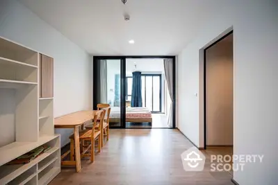 Spacious and modern apartment interior with open floor plan featuring a sleek wooden dining set, ample natural light, and a cozy bedroom visible through the sliding glass door.