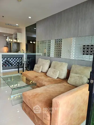 Modern living room with plush sofa and glass coffee table, open kitchen layout in background.