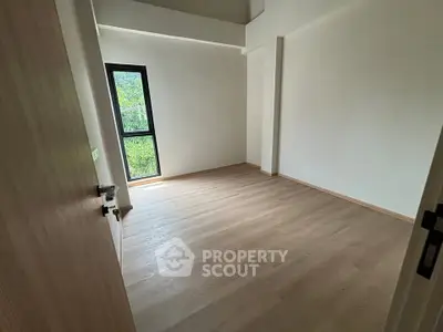 Spacious empty room with wooden flooring and large window offering lush green view.