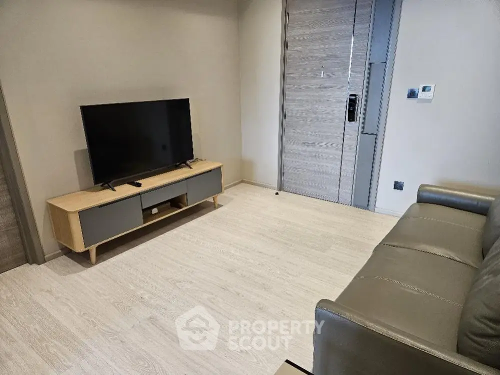Spacious and modern living room with sleek TV stand and comfortable couch, featuring light wood flooring and a secure entrance door.