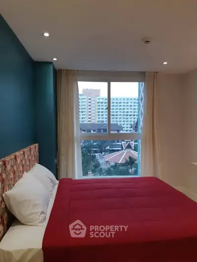 Modern bedroom with vibrant red bedding and city view from large window.