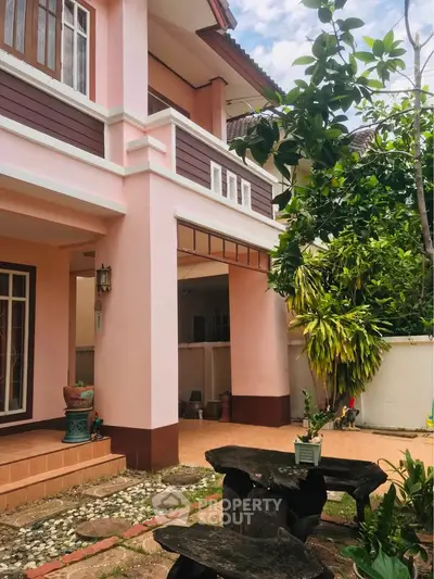 Charming two-story pink house with lush garden and outdoor seating area, perfect for family living.