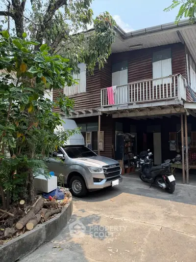 Charming two-story wooden house with balcony and driveway in lush green setting.