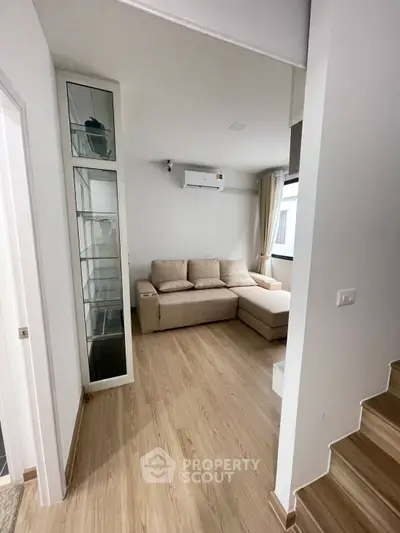 Modern living room with beige sofa and wooden flooring, featuring a glass display cabinet and air conditioning.