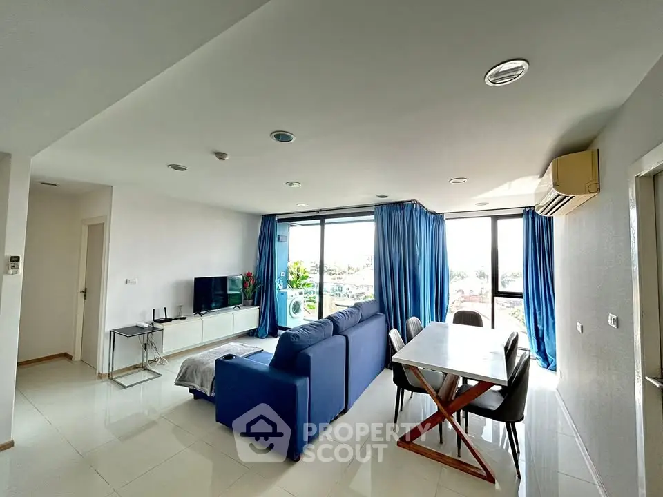 Spacious modern living room with blue sofa and dining area, featuring large windows and abundant natural light.