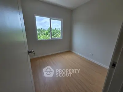 Bright empty room with wooden flooring and large window overlooking greenery.