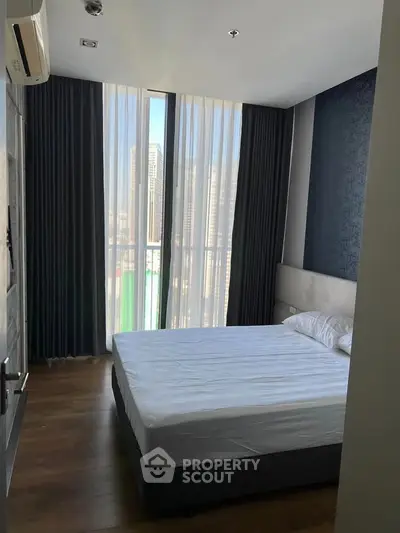 Modern bedroom with floor-to-ceiling windows and city view, featuring elegant curtains and wooden flooring.