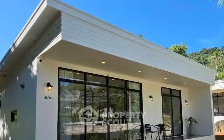 Modern single-story home with large windows and outdoor seating area, surrounded by lush greenery.