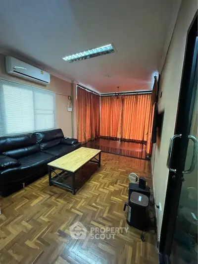 Cozy living room with parquet flooring and leather sofa, featuring large windows with orange curtains.