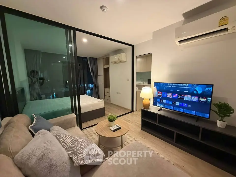 Modern living room with open layout, cozy sofa, and large TV, adjacent to a glass-walled bedroom.