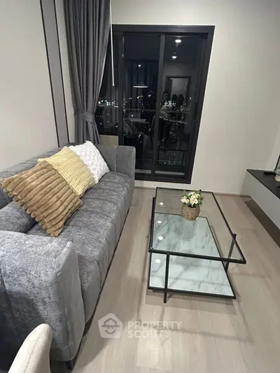 Modern living room with stylish gray sofa and glass coffee table, city view through large window.