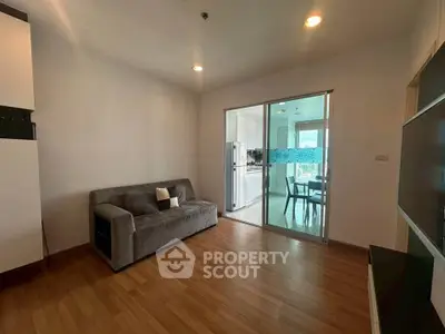 Modern living room with wooden flooring and sliding glass doors leading to a bright kitchen area.