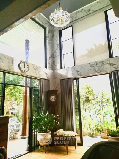 Luxurious living room with high ceilings, large windows, and elegant chandelier overlooking a lush garden.