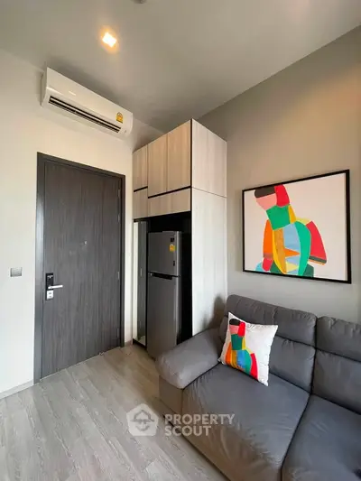 Modern living room with sleek furniture and vibrant artwork, featuring a compact kitchen area with built-in appliances.