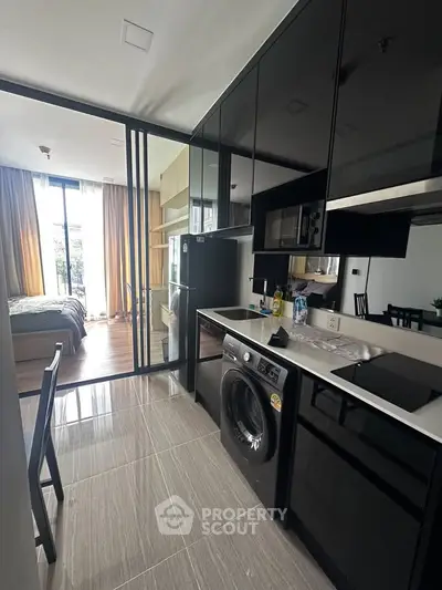 Modern apartment kitchen with sleek black cabinets and integrated appliances, leading to a cozy bedroom with natural light.