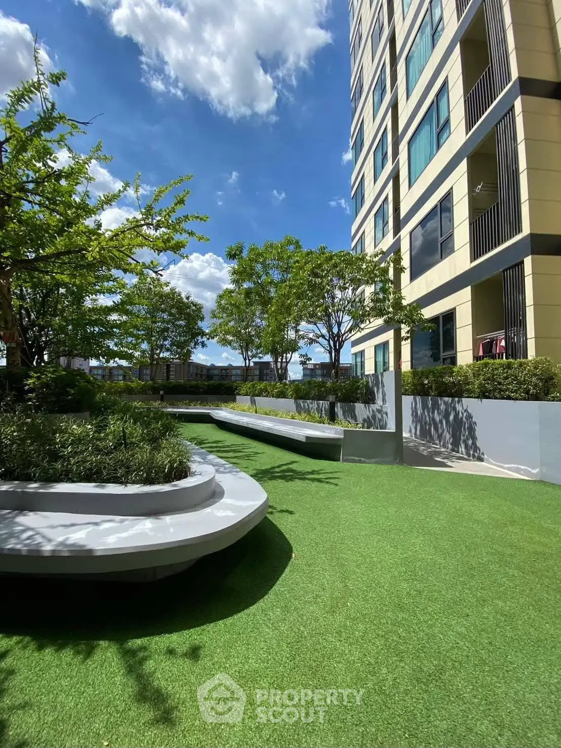 Modern building exterior with lush green garden and seating area under blue sky.
