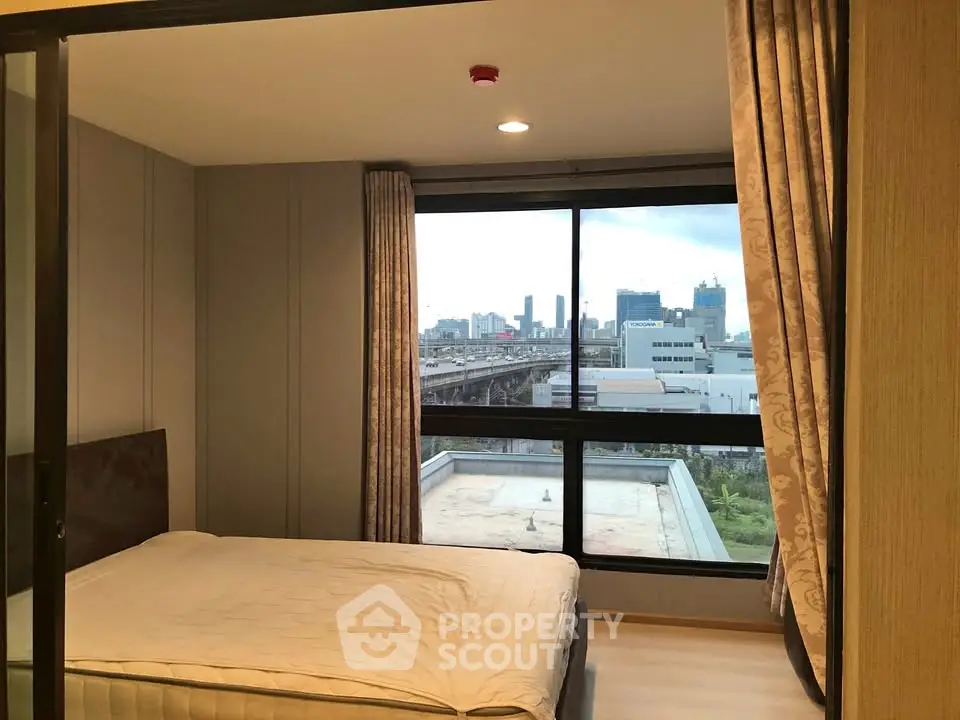 Modern bedroom with large window offering city skyline view, featuring elegant curtains and cozy bed.