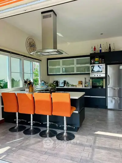 Modern kitchen with island, orange chairs, stainless steel appliances, and large windows.
