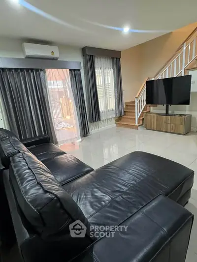 Spacious modern living room with sleek black leather sofa and large TV, featuring elegant staircase and natural light.