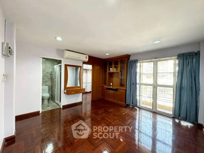 Spacious room with wooden flooring and built-in cabinets, featuring large windows and air conditioning.