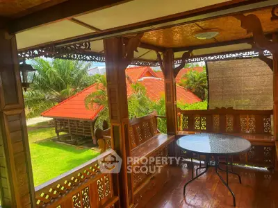 Charming wooden balcony with intricate carvings overlooking lush garden and red-roofed houses.