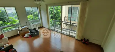 Spacious living room with large windows and balcony access, featuring wooden flooring and abundant natural light.